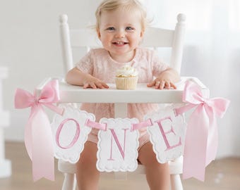 May include: A baby in a pink dress sits in a white high chair, smiling at the camera. A banner with the word "ONE" in pink letters hangs from the chair. A cupcake with white frosting sits on the tray. Pink bows adorn the sides of the banner.