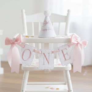 May include: A white high chair decorated for a first birthday. It features a banner that spells "ONE" in pink letters, a matching party hat with a pom-pom, and pink bows. Confetti is scattered on the chair.