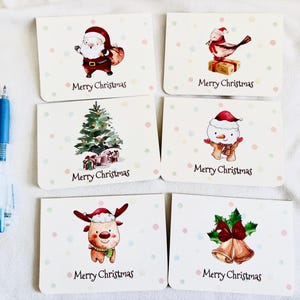 May include: A collection of six Christmas greeting cards with watercolor illustrations. Each card features a different festive design, including Santa Claus, a bird, a Christmas tree, a snowman, a reindeer, and bells. The words "Merry Christmas" are printed on each card.