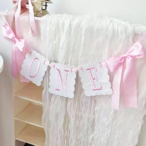 May include: A pink and white banner spelling "ONE" with scalloped edges and pink embroidered letters. The banner is strung with a pink ribbon and adorned with pink bows. The background is a white, draped fabric.