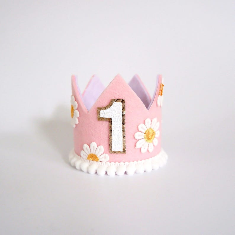 May include: A pink felt crown with a white number 1 outlined in gold glitter. The crown is decorated with white daisy flowers and a white pom-pom trim. The crown is designed for a first birthday celebration.