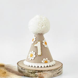 May include: A tan-colored birthday party hat with a white pom-pom on top. The hat is decorated with white daisies and the number "1" outlined in gold glitter. The base of the hat has a white pom-pom trim. The hat is displayed on a wooden slice.