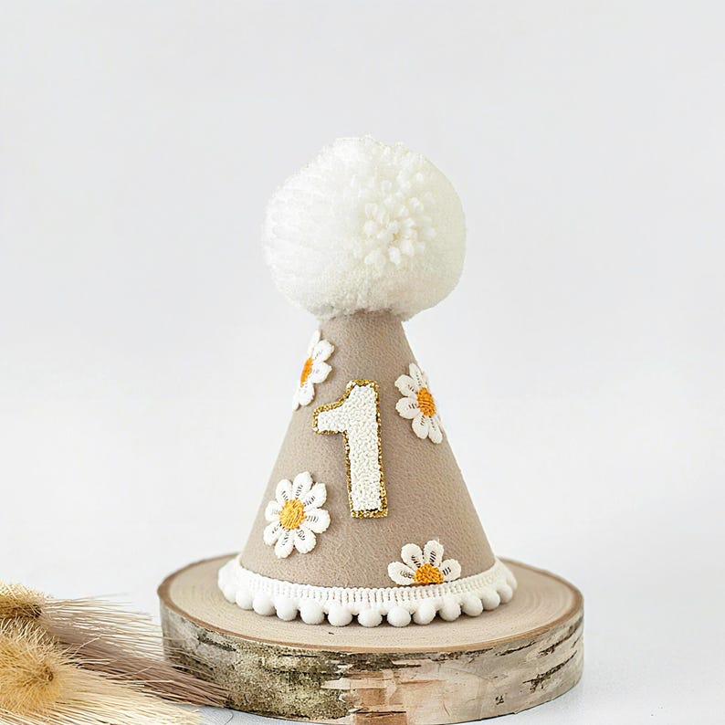 May include: A tan-colored birthday party hat with a white pom-pom on top. The hat is decorated with white daisies and the number "1" outlined in gold glitter. The base of the hat has a white pom-pom trim. The hat is displayed on a wooden slice.