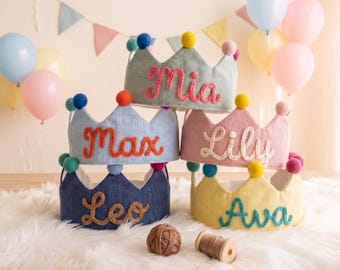 May include: Five colorful fabric crowns stacked on a white fluffy surface. Each crown is a different pastel color and has a name embroidered on it: Mia, Max, Lily, Leo, and Ava. The crowns are decorated with pom-poms.
