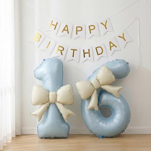 Pode incluir: Um conjunto de balões azuis claros com os números 1 e 6 com laços creme, perfeito para uma festa de aniversário. Uma faixa acima diz "HAPPY BIRTHDAY" em letras douradas.