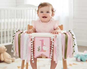 May include: A baby sits in a wooden high chair decorated with a pink and white banner. The banner features a number 1, a goose illustration, and various ribbons. The baby wears a pink outfit and smiles. A teddy bear and other toys are in the background.