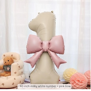 May include: A large, milky white number one balloon, approximately 40 inches tall, is adorned with a large pink bow. The balloon is a focal point, suitable for a first birthday celebration. The text at the bottom reads: 40 inch milky white number + pink bow.