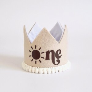 May include: A tan felt crown with the word "one" and a sun graphic in brown. The crown has a white interior and a white pom-pom trim at the base. A festive accessory for a first birthday celebration.