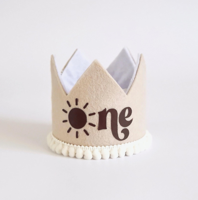 May include: A tan felt crown with the word "one" and a sun graphic in brown. The crown has a white interior and a white pom-pom trim at the base. A festive accessory for a first birthday celebration.