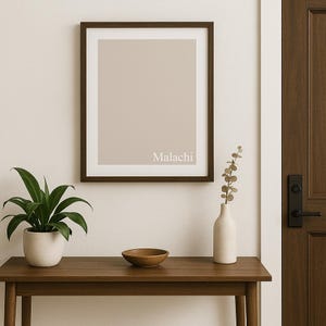 May include: A framed print with a beige background and the name "Malachi" in white lettering. The frame is dark brown. A small wooden table holds a potted plant, a wooden bowl, and a white vase with greenery.