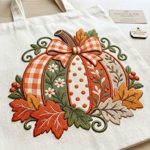 Puede incluir: Un bolso de tela color crema con un diseño de calabaza bordado. La calabaza tiene paneles de cuadros naranjas, naranja sólido y lunares blancos, decorada con un lazo de cuadros, flores blancas, enredaderas verdes y hojas de otoño naranjas.