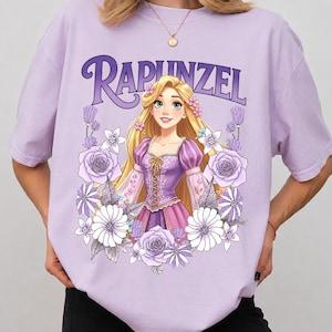 Puede incluir: Una camiseta lila claro con la palabra "Rapunzel" en una fuente estilizada sobre una ilustraci&oacute;n de dibujos animados de una princesa rubia con un vestido morado, rodeada de flores. La camiseta es de material suave.