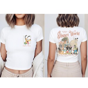May include: White crop top featuring Pluto with a green floral design and the text "Custom Name" on the front. The back of the shirt displays "Florida Disney World" with various Disney characters and park landmarks.