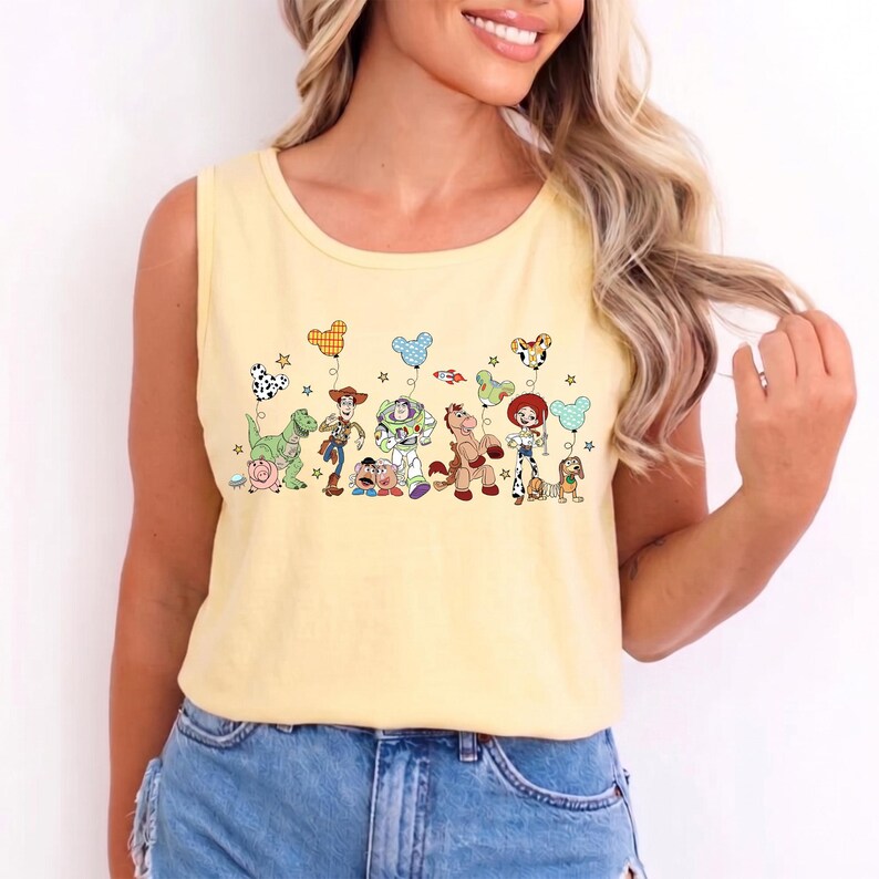 May include: A yellow tank top featuring a colorful graphic of cartoon characters and balloons. The design includes a dinosaur, a cowboy, a space ranger, a horse, and other characters. The balloons are in various shapes and patterns.