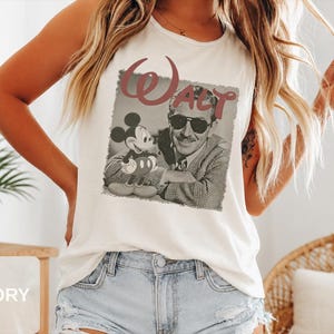 Peut inclure: Un d&eacute;bardeur sans manches ivoire avec une impression graphique de style vintage repr&eacute;sentant Walt Disney et Mickey Mouse. L'image en noir et blanc montre Walt Disney portant des lunettes de soleil et une veste &agrave; motifs, avec le mot WALT en grandes lettres rouges au-dessus.