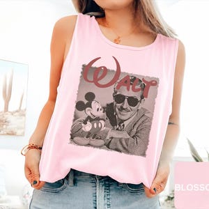 Peut inclure: Un d&eacute;bardeur rose clair sans manches avec une impression graphique de Walt Disney portant des lunettes de soleil &agrave; c&ocirc;t&eacute; de Mickey Mouse. Le mot Walt est imprim&eacute; dans une police rouge fonc&eacute; stylis&eacute;e au-dessus de l'image photographique en noir et blanc.