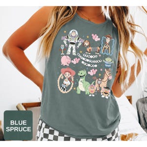 May include: A blue spruce sleeveless shirt featuring a graphic print of Toy Story characters including Buzz Lightyear, Woody, Jessie, Rex, Slinky Dog, Hamm, and the Aliens. The text "YOU'VE GOT A FRIEND IN ME" is printed in the center with pink balloons.
