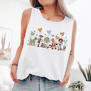 May include: White tank top featuring a colorful graphic of animated characters. The design includes a green dinosaur, a cowboy, a space ranger, and other characters. The tank top is sleeveless and made of a soft material.