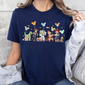 K&ouml;nnte beinhalten: Ein marineblaues Kurzarm-T-Shirt mit einem Grafikdruck von Toy Story-Figuren wie Woody, Buzz Lightyear, Jessie, Rex, Hamm, Slinky Dog und Mr. Potato Head, die jeweils einen gemusterten, Micky-Maus-f&ouml;rmigen Luftballon halten.