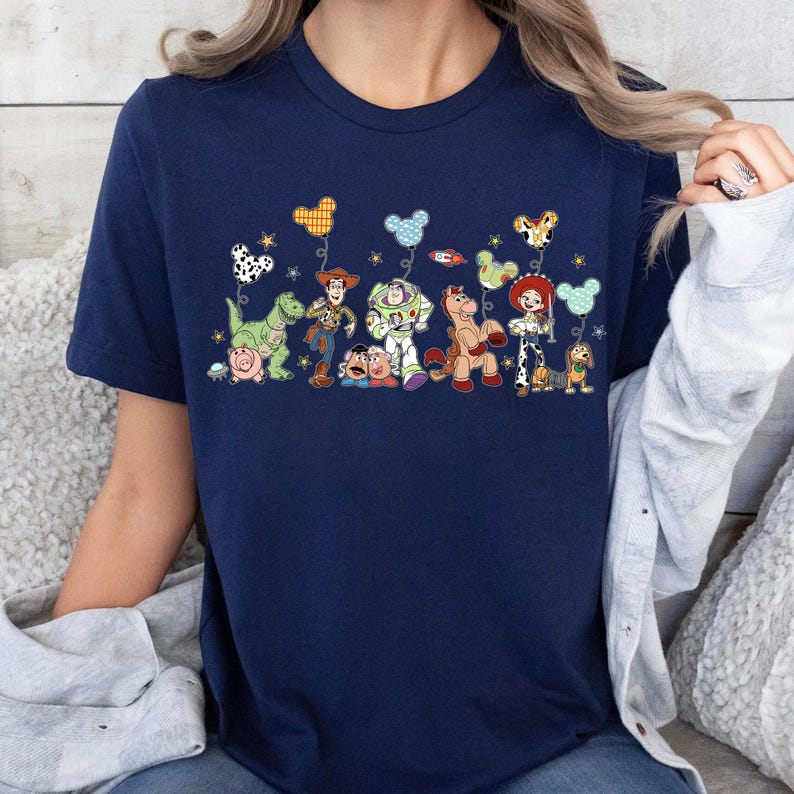 K&ouml;nnte beinhalten: Ein marineblaues Kurzarm-T-Shirt mit einem Grafikdruck von Toy Story-Figuren wie Woody, Buzz Lightyear, Jessie, Rex, Hamm, Slinky Dog und Mr. Potato Head, die jeweils einen gemusterten, Micky-Maus-f&ouml;rmigen Luftballon halten.