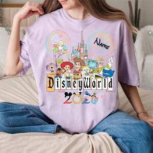Peut inclure: T-shirt lilas avec un motif Disney World. Le motif comprend un ch&acirc;teau, des personnages Disney, les mots "Disneyworld 2020" et un espace pour un nom. Le t-shirt est de style d&eacute;contract&eacute;.