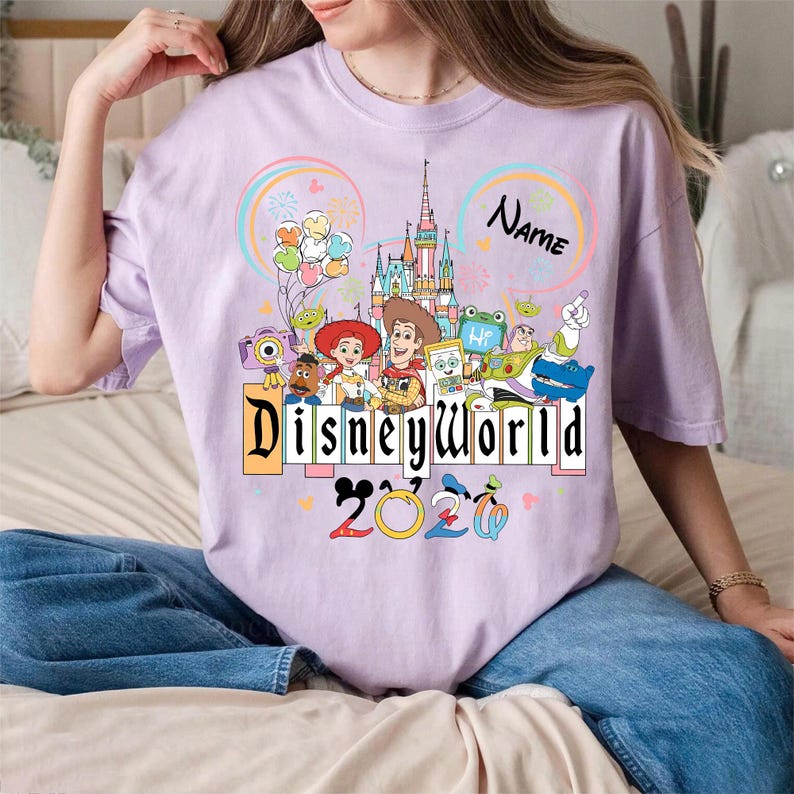 Peut inclure: T-shirt lilas avec un motif Disney World. Le motif comprend un ch&acirc;teau, des personnages Disney, les mots "Disneyworld 2020" et un espace pour un nom. Le t-shirt est de style d&eacute;contract&eacute;.
