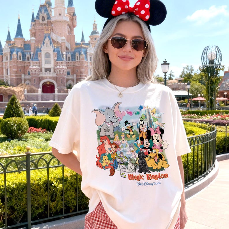 K&ouml;nnte beinhalten: Cremefarbenes T-Shirt mit einer bunten Grafik von Disney-Figuren, darunter Dumbo, Mickey Mouse und Arielle. Der Text "Magic Kingdom" ist unter den Figuren aufgedruckt. Die Person tr&auml;gt eine Sonnenbrille und ein Minnie Mouse Ohren-Stirnband.