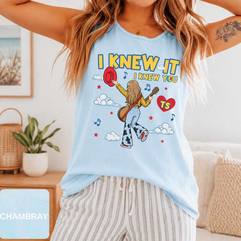 May include: A light blue sleeveless tank top featuring a graphic illustration of a person with long hair holding a red hat and a guitar. The text reads I KNEW IT I KNEW YOU with a red heart containing the letters TS, surrounded by clouds and stars.