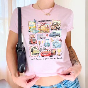 May include: Light pink t-shirt featuring a colorful graphic of cartoon cars and the text "Radiator Springs" and "I eat lasers for breakfast". The design includes various car illustrations and decorative elements.