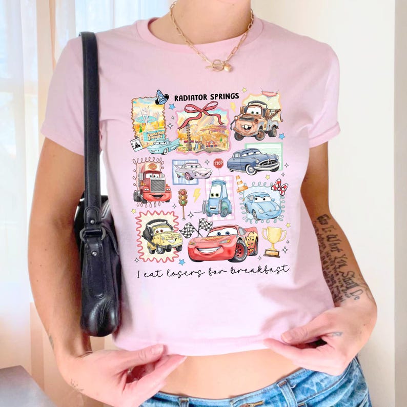 May include: Light pink t-shirt featuring a colorful graphic of cartoon cars and the text "Radiator Springs" and "I eat lasers for breakfast". The design includes various car illustrations and decorative elements.