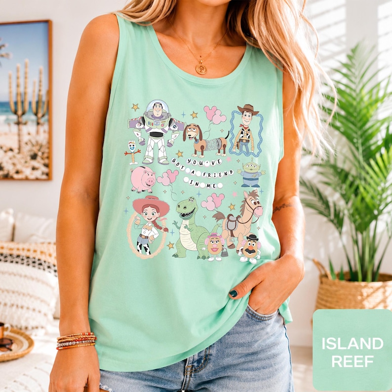 May include: A light mint green sleeveless tank top featuring a colorful graphic print of various Toy Story characters including Buzz Lightyear, Woody, Jessie, and Rex, with the text "YOU'VE GOT A FRIEND IN ME" centered among the illustrated figures.