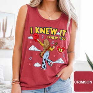 May include: A crimson sleeveless tank top featuring a graphic illustration of a person with long hair holding a guitar and a red hat. The text reads I KNEW IT I KNEW YOU with a heart containing the letters TS, surrounded by clouds, stars, and music notes.