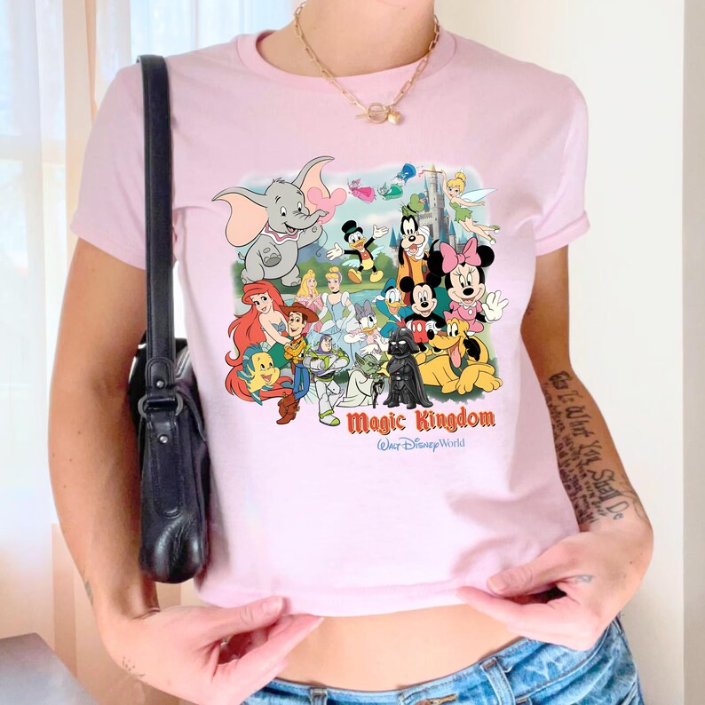 K&ouml;nnte beinhalten: Hellrosa T-Shirt mit einem farbenfrohen Aufdruck verschiedener Disney-Figuren, darunter Mickey Mouse, Minnie Mouse und Dumbo. Der Text "Magic Kingdom Walt Disney World" ist unter den Figuren aufgedruckt.
