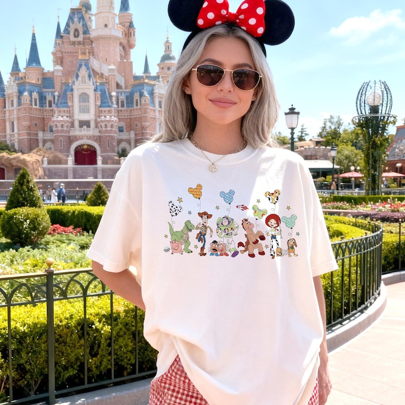 K&ouml;nnte beinhalten: Ein wei&szlig;es T-Shirt mit einem Grafikdruck verschiedener animierter Spielzeugfiguren, die in einer Reihe stehen und jeweils einen bunten, Mickey-Maus-f&ouml;rmigen Luftballon halten. Das Design enth&auml;lt kleine Sternakzente um die Figuren.