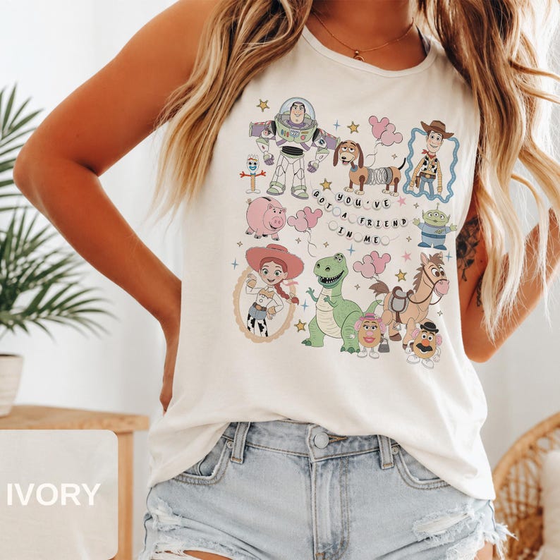 May include: An ivory sleeveless tank top featuring a colorful graphic print of various animated characters including a space ranger, a cowboy, a cowgirl, a dinosaur, a pig, and a dog. The text "YOU'VE GOT A FRIEND IN ME" is printed in the center.
