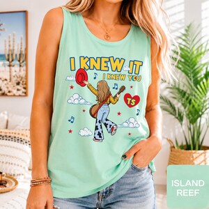 May include: A light green sleeveless tank top featuring a graphic illustration of a person with long hair holding a red hat and a guitar. The text reads I KNEW IT I KNEW YOU with a red heart containing the letters TS, blue musical notes, and stars.