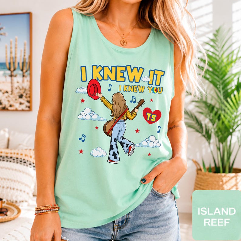May include: A light green sleeveless tank top featuring a graphic illustration of a person with long hair holding a red hat and a guitar. The text reads I KNEW IT I KNEW YOU with a red heart containing the letters TS, blue musical notes, and stars.