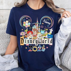 Peut inclure: T-shirt bleu marine avec un motif Disney World color&eacute;. Le motif comprend des personnages Disney, un ch&acirc;teau, des feux d'artifice et le texte "Disneyworld 2020". Un espace est disponible pour un nom.