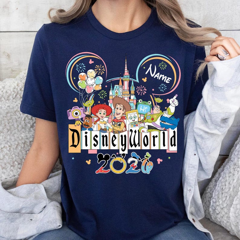 Peut inclure: T-shirt bleu marine avec un motif Disney World color&eacute;. Le motif comprend des personnages Disney, un ch&acirc;teau, des feux d'artifice et le texte "Disneyworld 2020". Un espace est disponible pour un nom.