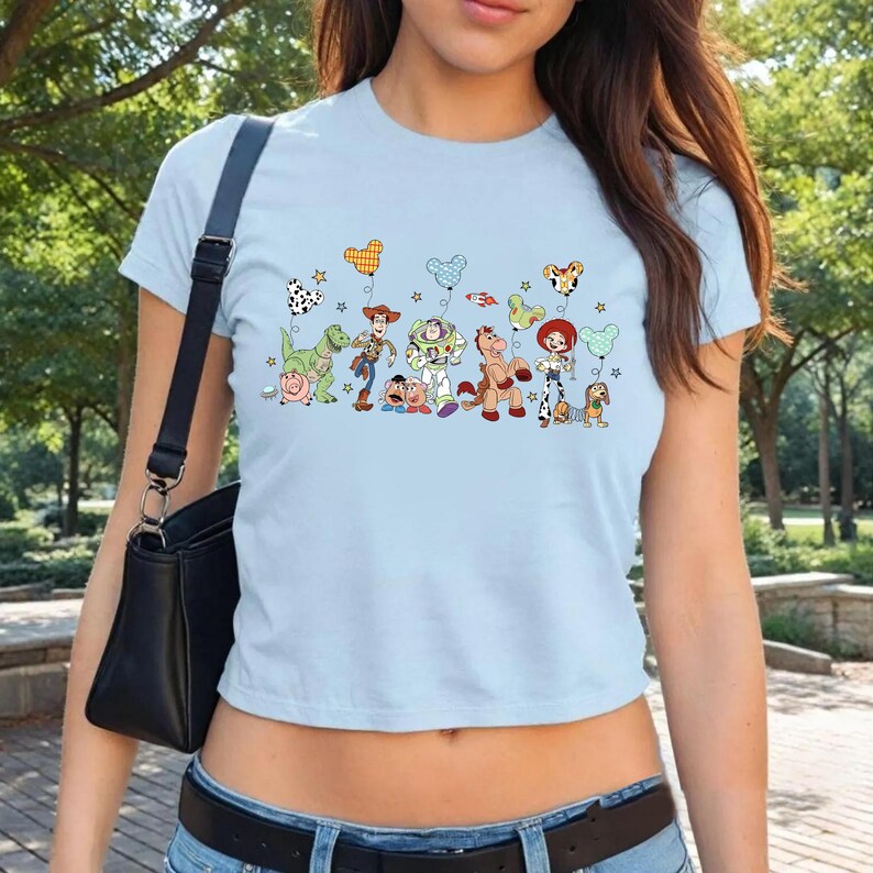 K&ouml;nnte beinhalten: Ein hellblaues T-Shirt mit kurzen &Auml;rmeln und einem Grafikdruck verschiedener Toy Story-Figuren, darunter Woody, Buzz Lightyear und Jessie, die jeweils einen Mickey-Maus-f&ouml;rmigen Ballon halten, erg&auml;nzt durch kleine Sterne und eine Rakete.
