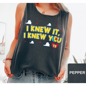 May include: A dark grey sleeveless tank top featuring the yellow text I KNEW IT, I KNEW YOU with a colourful ball graphic replacing the letter O, a red heart with the letters TS, and small white cloud and red star icons printed on the front.