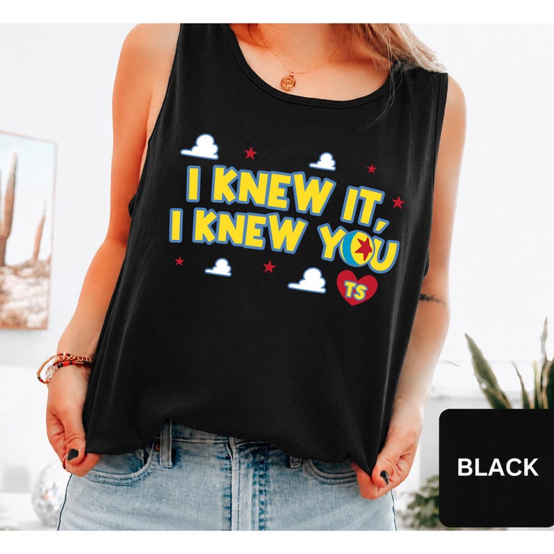 May include: A black sleeveless tank top featuring the yellow text I KNEW IT, I KNEW YOU with a colourful ball graphic replacing the letter O. The design includes small white clouds and red stars, plus a red heart with the letters TS inside.