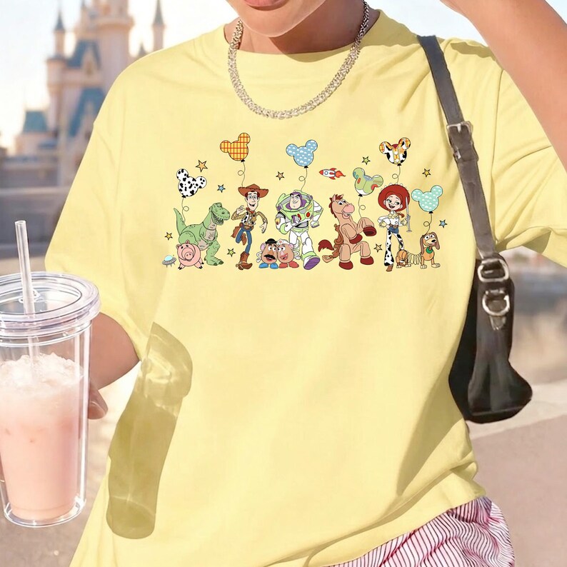 K&ouml;nnte beinhalten: Ein hellgelbes T-Shirt mit einem Grafikdruck verschiedener Zeichentrickfiguren, darunter ein Cowboy, ein Weltraum-Ranger, ein Cowgirl, ein Dinosaurier, ein Schwein und ein Pferd, die alle Luftballons in Mausohrform und kleine Sterne halten.