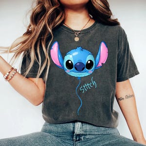 May include: A charcoal gray short-sleeve t-shirt featuring a graphic of a blue character head shaped like a balloon with large black eyes and pink inner ears. A thin blue string hangs from the character with the word Stitch written in blue script.