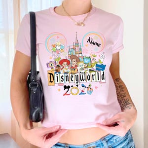 Peut inclure: T-shirt rose clair avec un motif Disney World color&eacute;. Le motif comprend des personnages de dessins anim&eacute;s, un ch&acirc;teau et les mots "Disneyworld 2020". Le t-shirt a un col rond et des manches courtes.