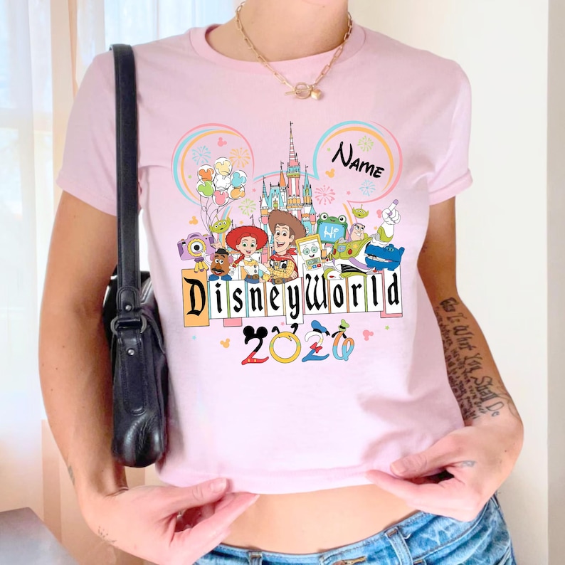 Peut inclure: T-shirt rose clair avec un motif Disney World color&eacute;. Le motif comprend des personnages de dessins anim&eacute;s, un ch&acirc;teau et les mots "Disneyworld 2020". Le t-shirt a un col rond et des manches courtes.