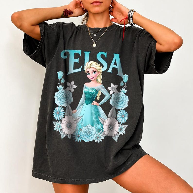 Puede incluir: Camiseta negra con el nombre "ELSA" en turquesa sobre una imagen de dibujos animados de una mujer rubia con un vestido turquesa. La imagen est&aacute; enmarcada por flores azules y blancas.