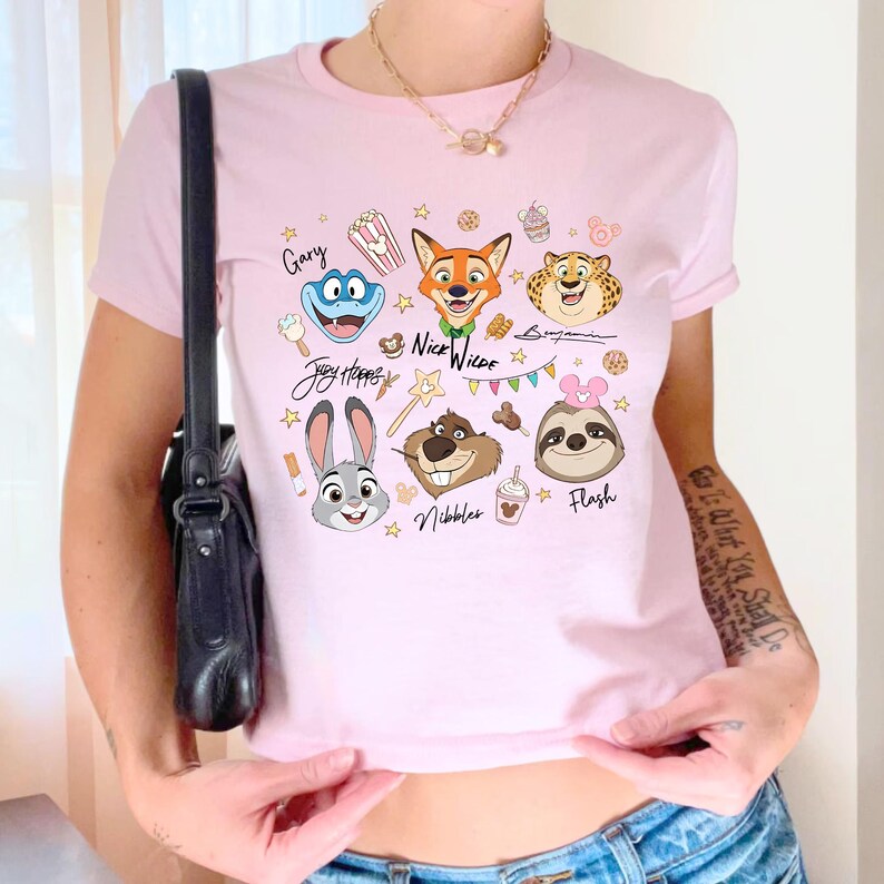 Pu&ograve; includere: T-shirt rosa chiaro con illustrazioni di personaggi dei cartoni animati e i nomi Gary, Judy Hopps, Nick Wilde, Benjamin, Nibbles e Flash. Il design include stelle, cibo e uno striscione. La maglietta &egrave; a maniche corte.