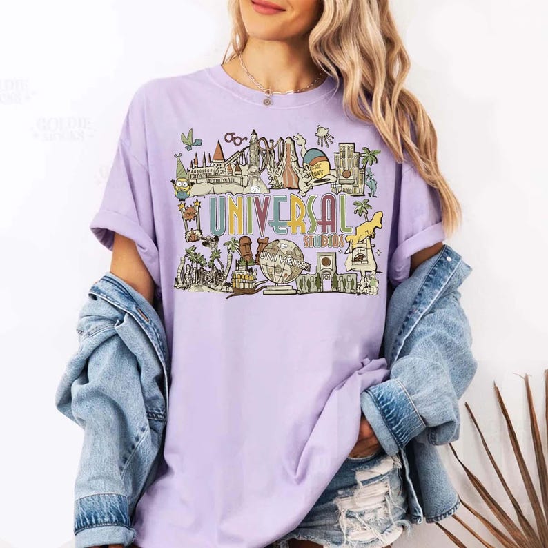 May include: A lilac t-shirt with a colourful graphic design featuring the words "UNIVERSAL STUDIOS". The design includes illustrations of landmarks and characters associated with Universal Studios.