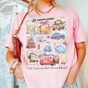 May include: Pink t-shirt featuring a collage of cartoon cars and the text "Radiator Springs." The design includes various car characters, a trophy, and the phrase "I eat losers for breakfast" in black script.